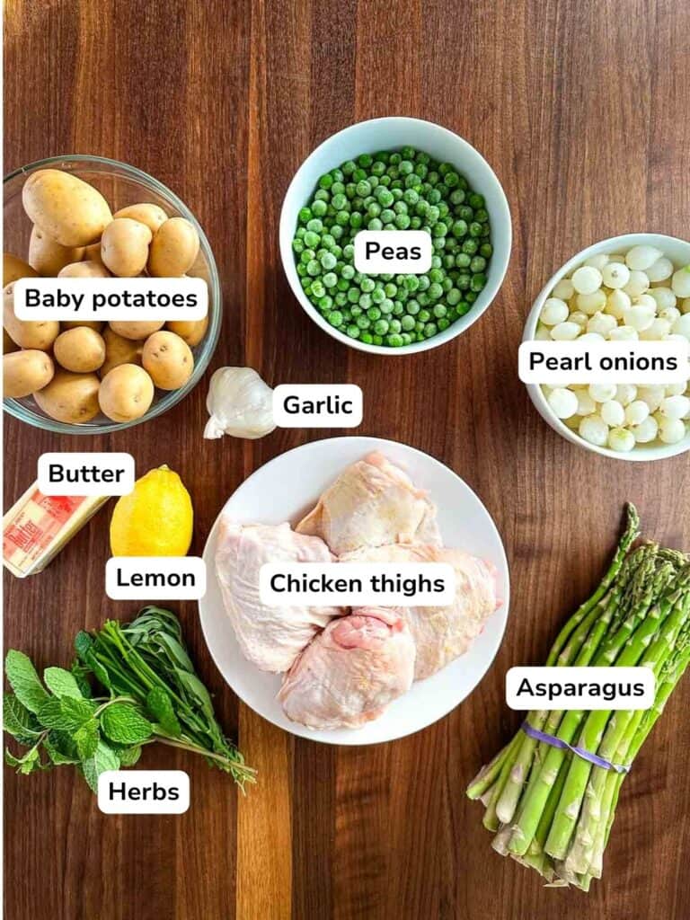 An over head photo of ingredients required to make the recipe: a bowl of potatoes, a bowl of peas, a bowl of pearl onions, garlic, lemon, butter, fresh herbs, a bunch of asparagus and a plate of chicken thighs.