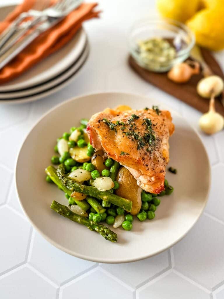 A cream colored with plate with roasted chicken, asparagus and peas next to a bowl of herb butter, two lemons and some pearl onions and a stack of plates and flatware with orange napkins.