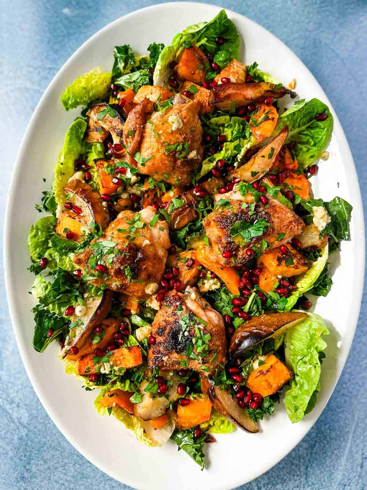 A white platter with dressed greens piled with roasted chicken, squash, pears, blue cheese, pomegranate seeds and fresh parsley.