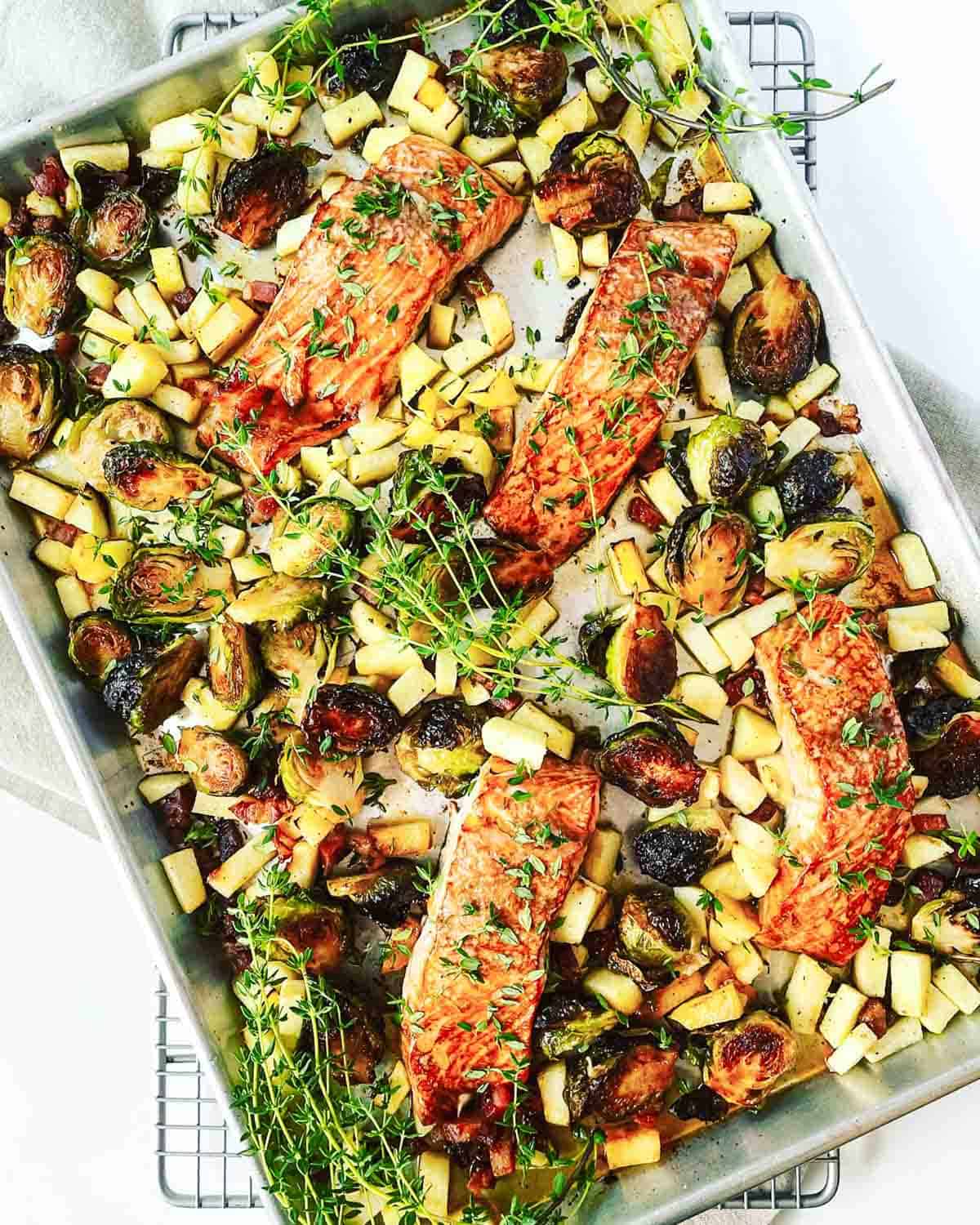 A sheet pan filled with roasted salmon filets, brussels sprouts, apples, pancetta and fresh thyme.