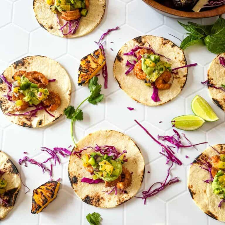 A white hex tile background with several tortillas layered with red cabbage, roasted shrimp, pineapple salsa and avocado sauce next to lime wedges and a wooden bowl full of avocados.
