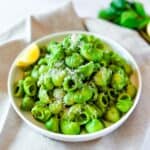 A white plate on a linen cloth with bright green pasta and a lemon wedge.