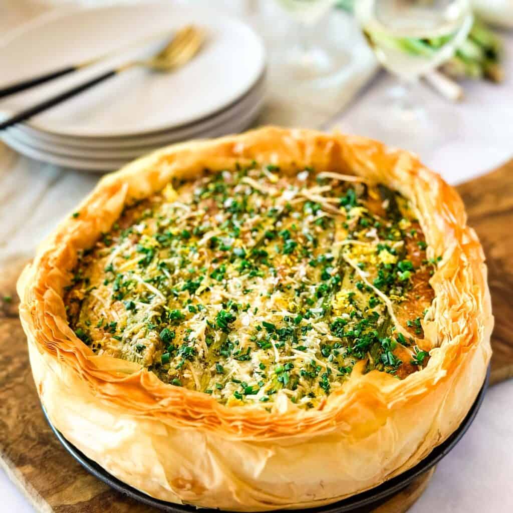 Asparagus and leek tart with phyllo dough on a cutting board next to a stack of plates with silverware and two glasses of white wine in the background.