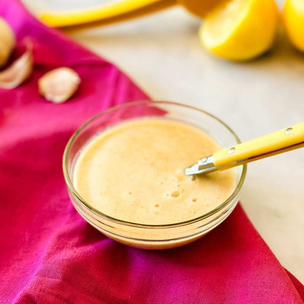 A small glass bowl of lemon brown butter vinaigrette on a bright pink napkin next to lemons.