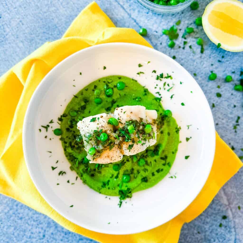 A blue background and bright yellow napkin with a white plate filled with one pan halibut with minted pea puree next to half a lemon and some scattered peas.