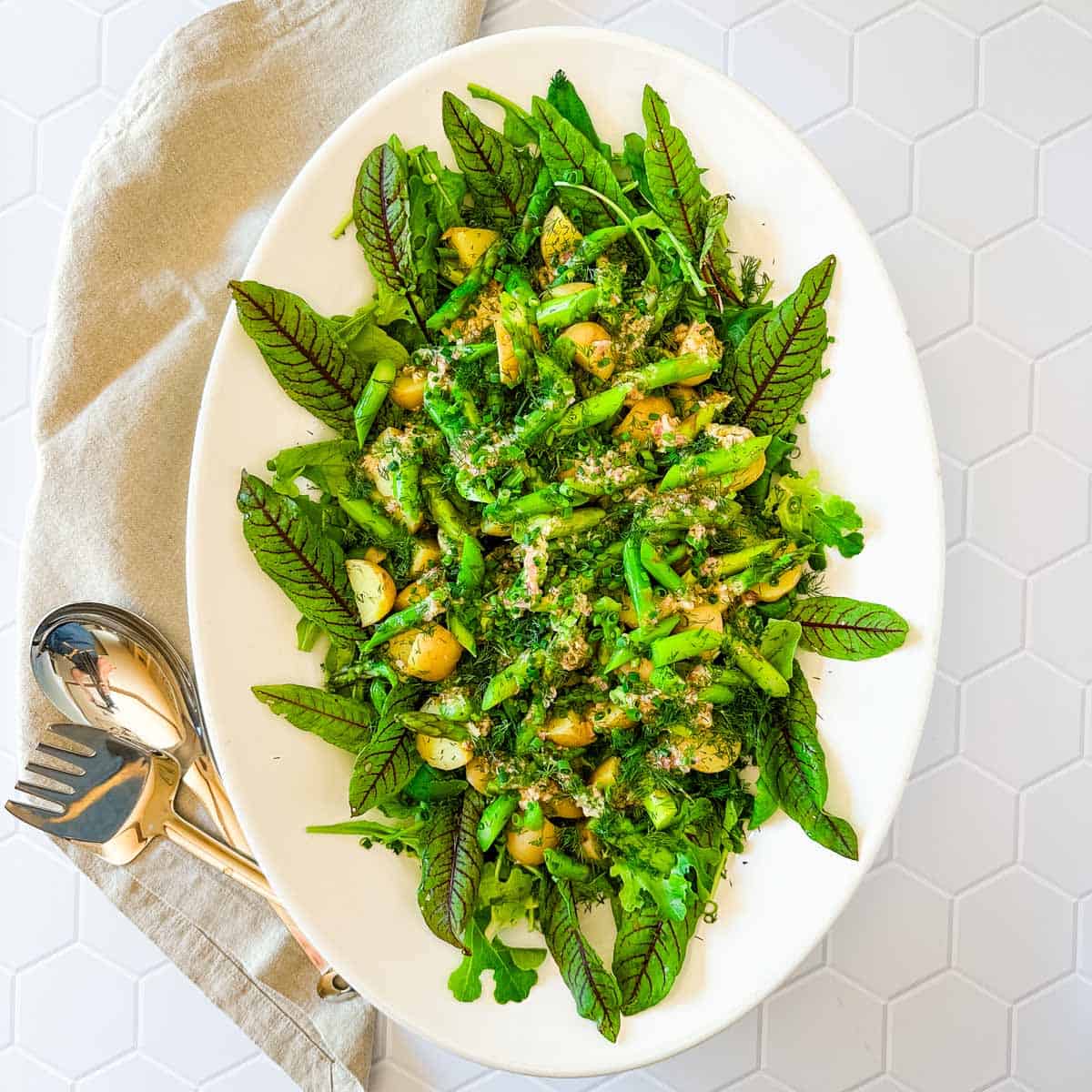 A large white oval platter with potato and asparagus salad on a linen napkin with serving utensils.