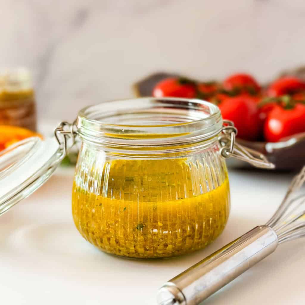A glass jar filled with orange mustard vinaigrette next to a whisk and a bowl of red tomatoes.