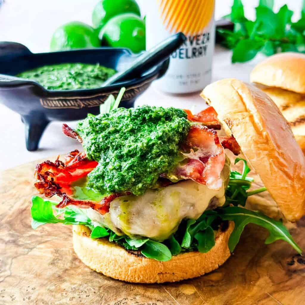 A front view of grilled lamb burger with bacon and mint-jalapeno chimichurri on a brioche bun next to a bowl of sauce and a can of beer.