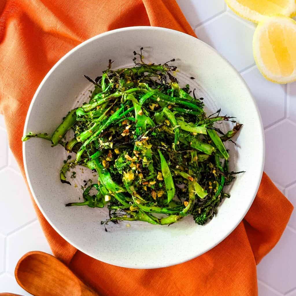 An off white bowl of grilled broccolini on an orange napkin next to lemon slices and wooden utensils.