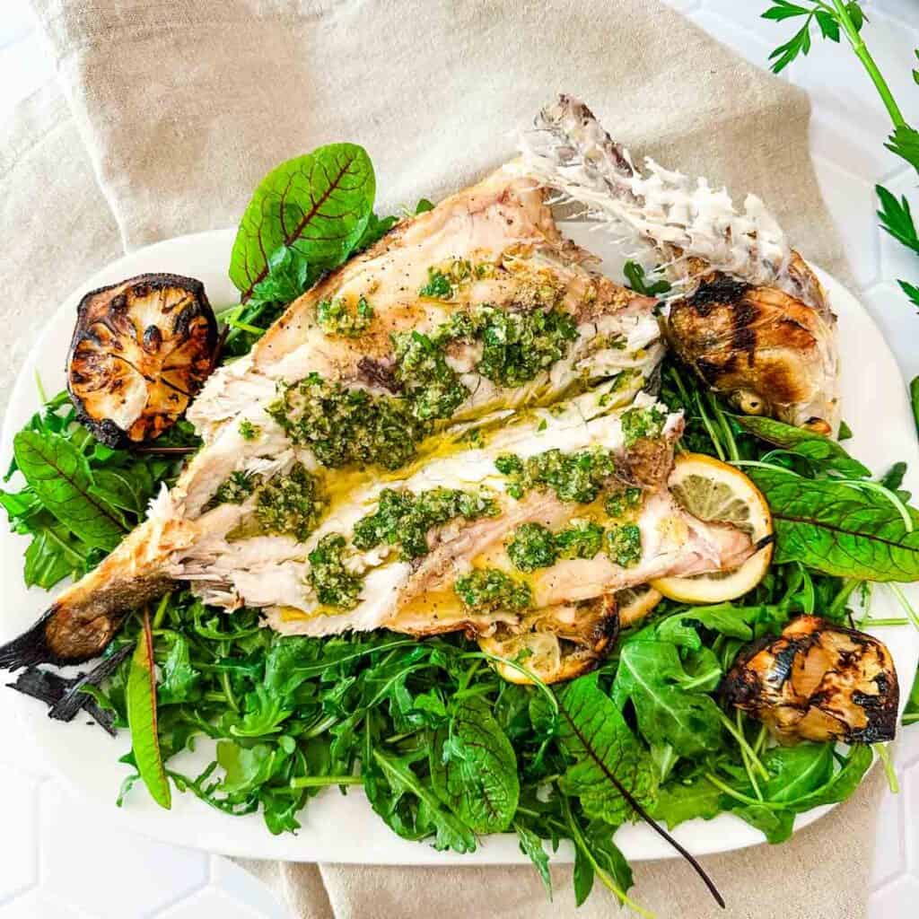 A white serving platter with arugula and whole grilled branzino and grilled lemons.