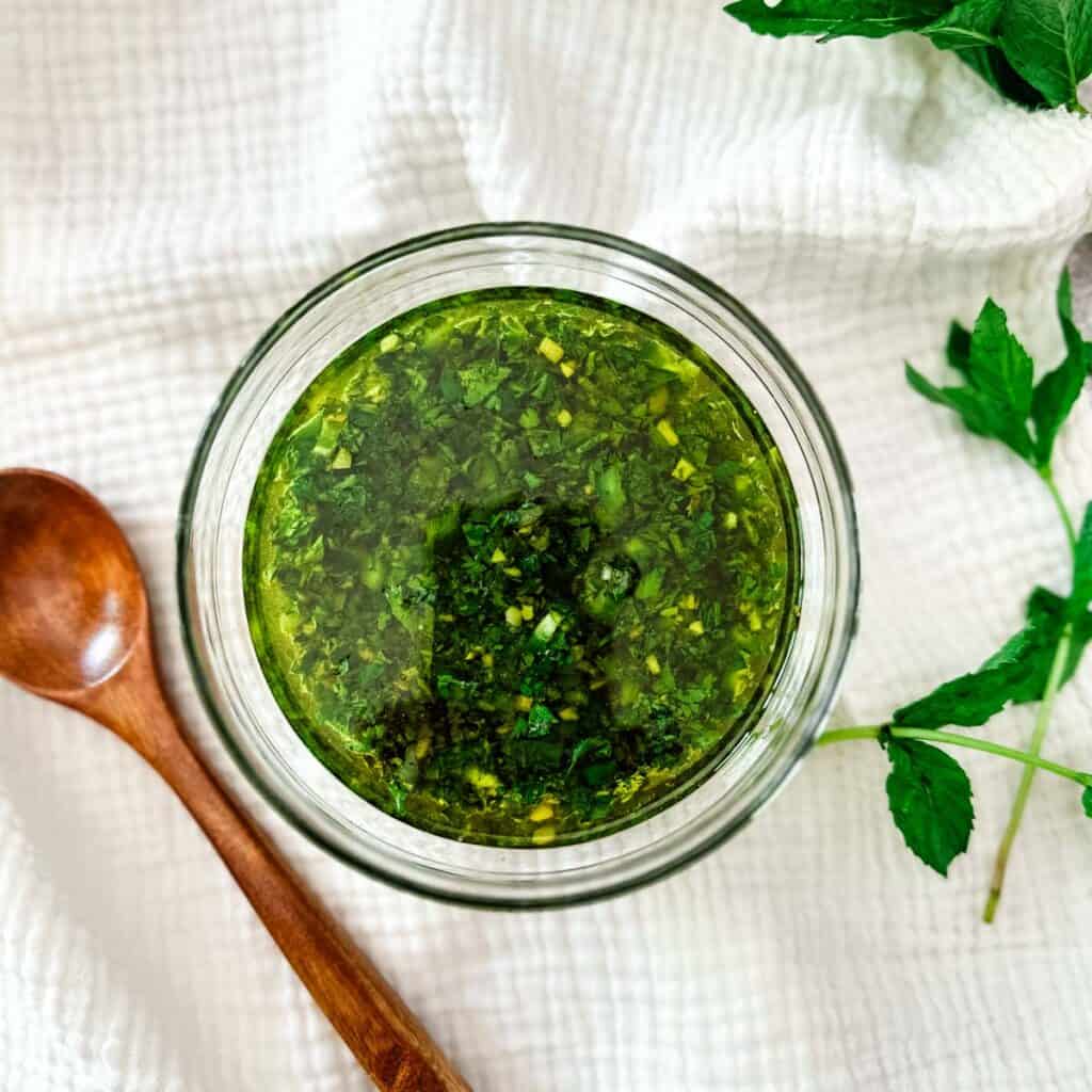 A glass bowl of mint-jalapeno chimichurri next to a small wooden spoon and sprigs of mint.