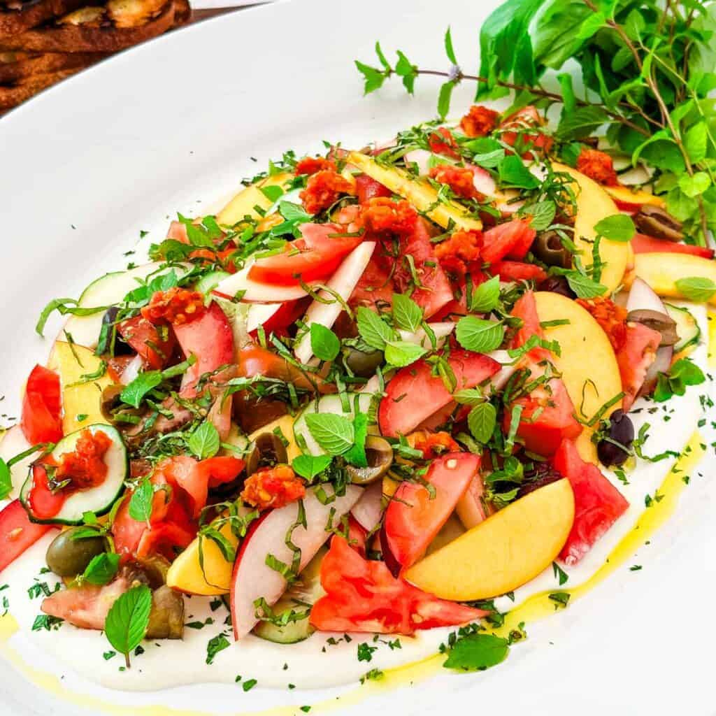 An oval white platter with whipped feta with tomatoes, peaches and grilled bread.