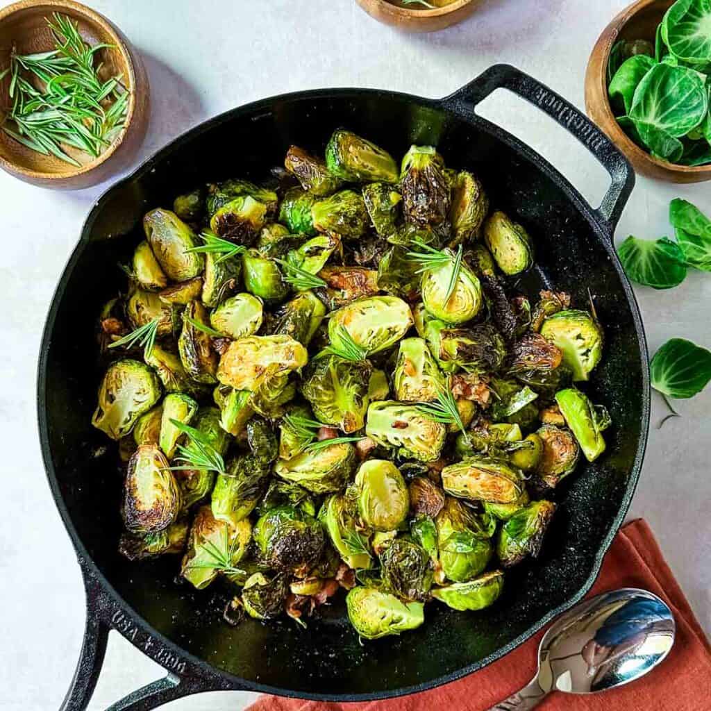 An overhead view of a cast iron skillet filled with brussels sprouts with pancetta, rosemary and garlic next to wooden bowls of rosemary and an orange napkin.