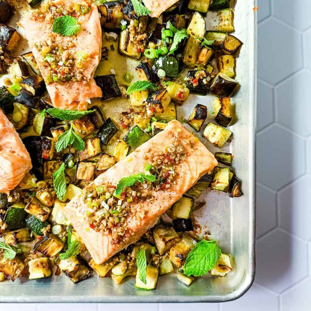 An overhead view of a corner of a sheet pan with salmon, eggplant, zucchini and chili crisp.