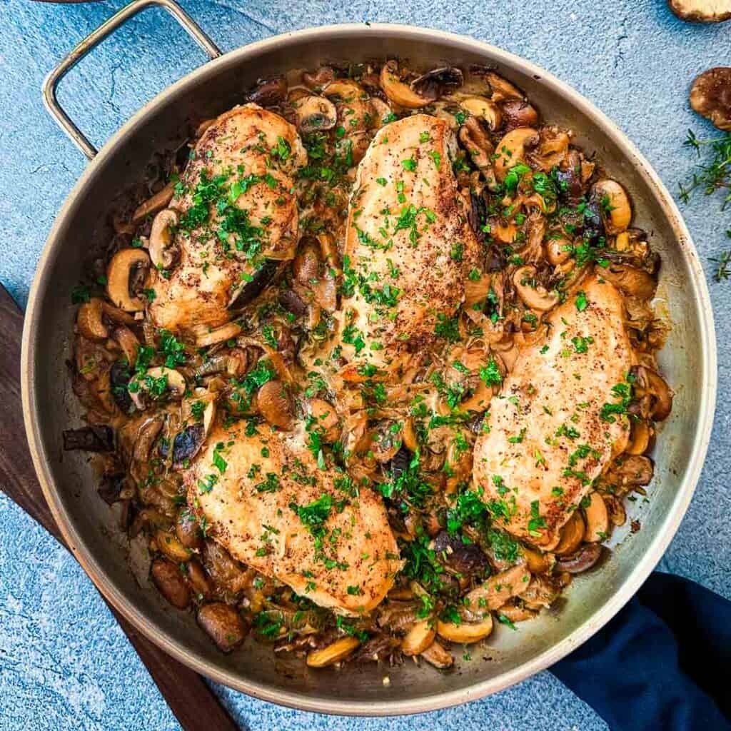 An overview of a skillet with chicken, mushrooms and caramelized onions on a blue background next to thyme sprigs and a wooden spoon.