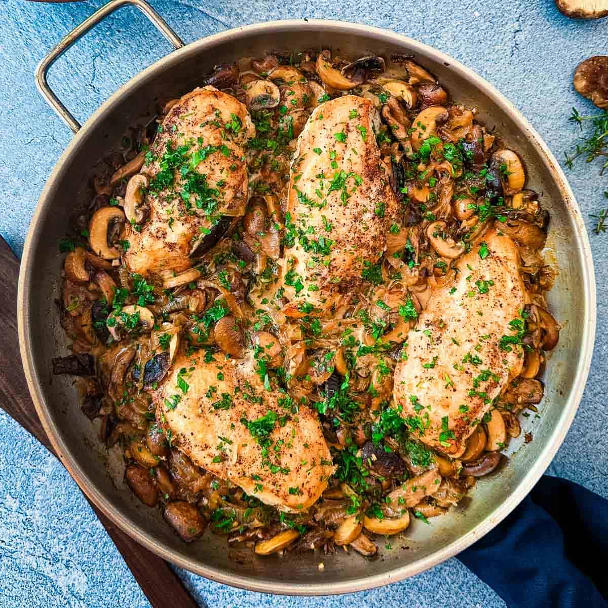 A saute pan filled with chicken, mushrooms, and caramelized onions on a blue background.