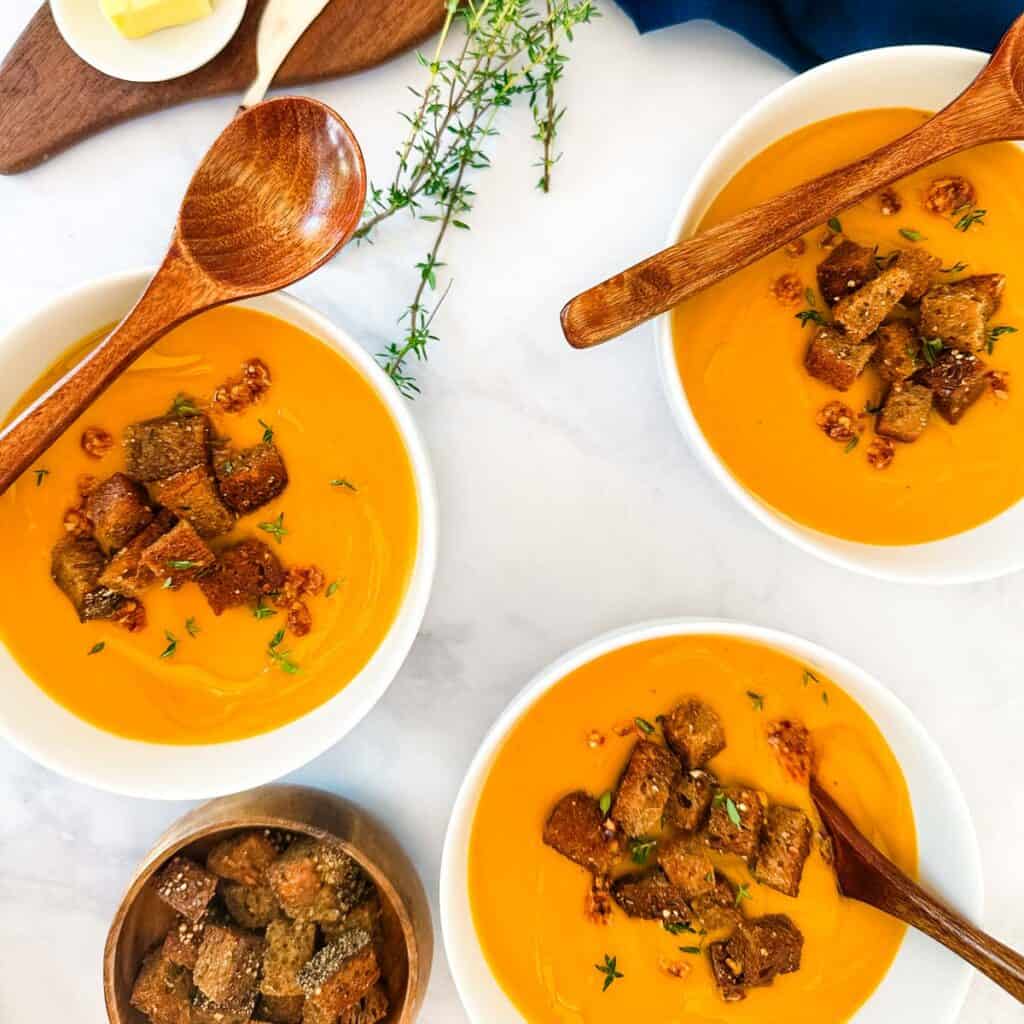 An overhead view of three white bowls of carrot lentil soup next to a stack of bread and butter and a navy blue napkin.