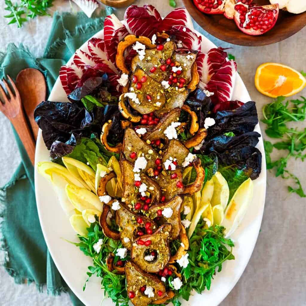 An overhead view of a roasted pear salad on a white oval platter next to a green napkin with wooden serving utensils, a cut orange, a split open pomegranate.