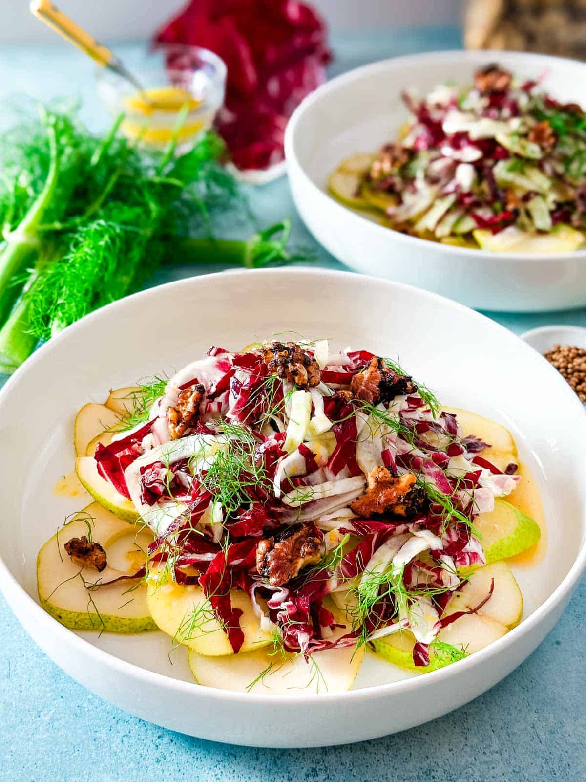 Two shallow white bowls with pear salad next to fennel fronds, radicchio leaves, and a glass bowl of lemon vinaigrette.