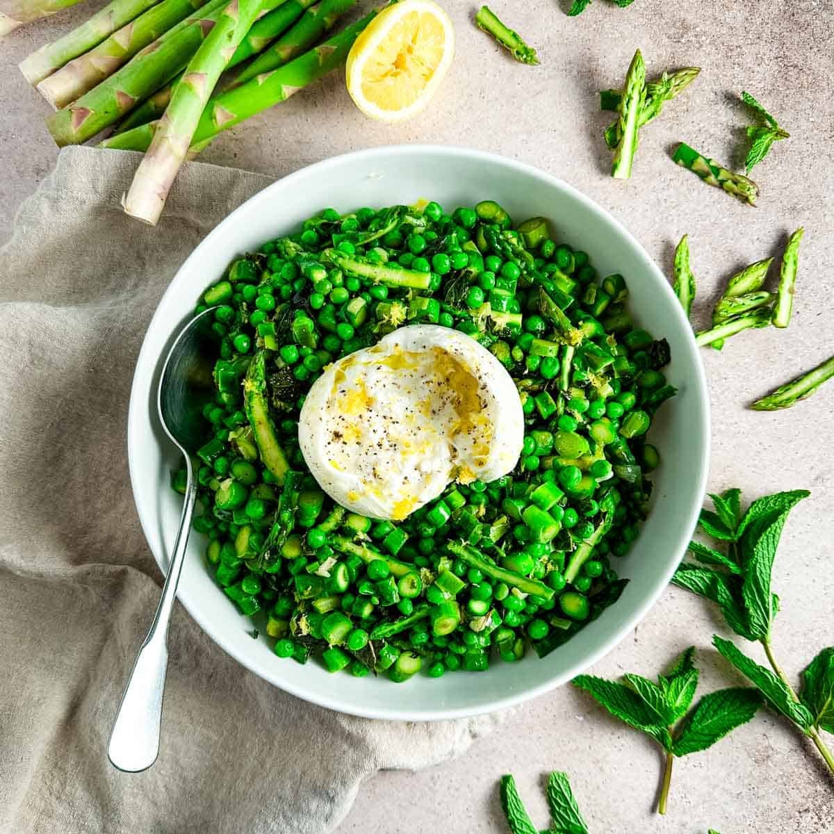 A white shallow bowl of sautéed asparagus and peas next to mint sprigs and asparagus spears.
