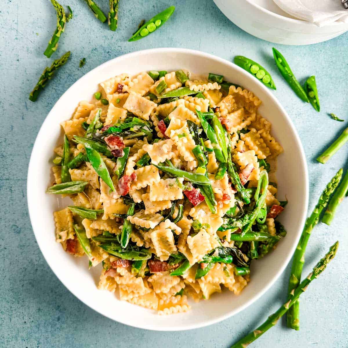 A white shallow bowl of snap pea and asparagus pasta with bacon on a light blue background.