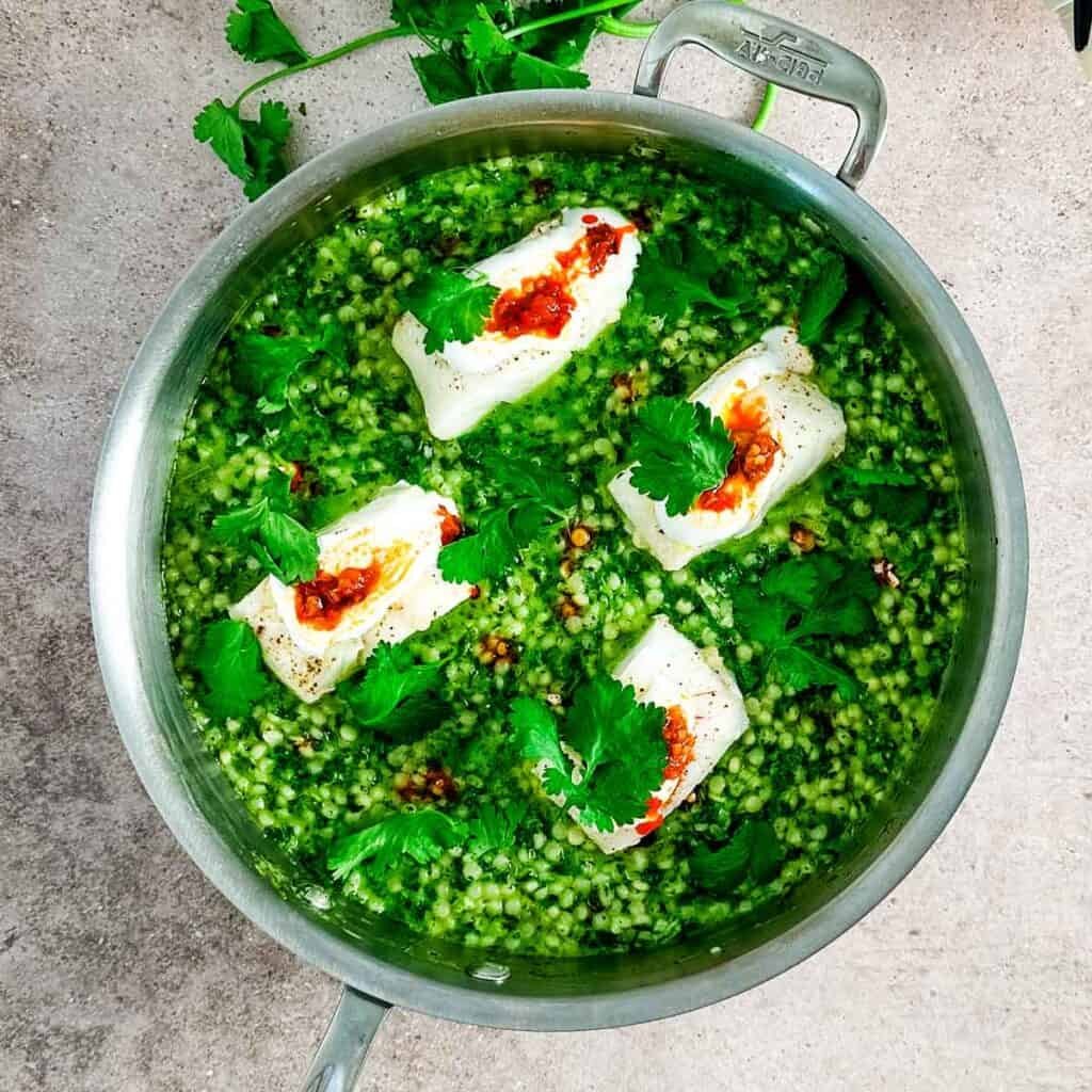 A large saute pan with green herby couscous with cod fillets on top on a neutral background.