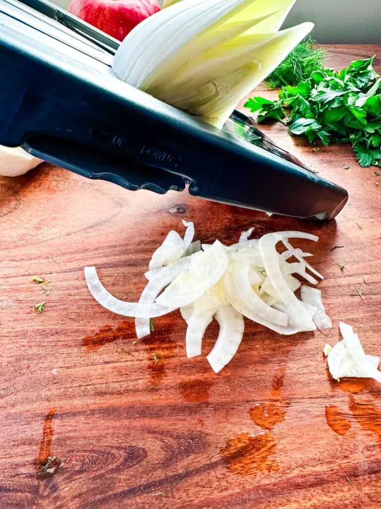 Halved fennel bulbs being thinly sliced on a mandolin on a cutting board.