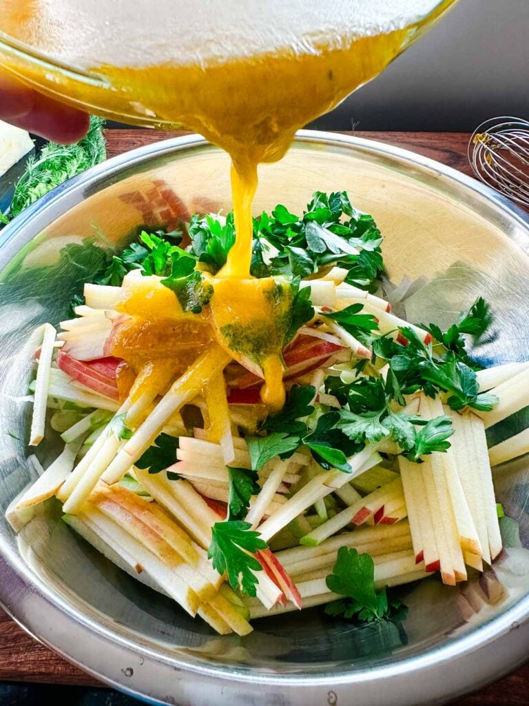 Ginger vinaigrette being poured onto apple fennel slaw in a large stainless steel bowl.