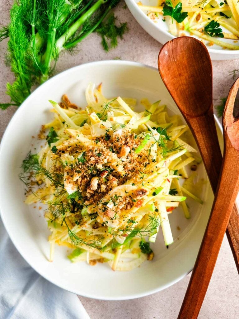 Two white shallow bowls with apple fennel slaw with wooden serving utensils on a neutral background.