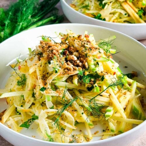 A white shallow bowl with apple fennel slaw in a high pile next to fennel fronds.