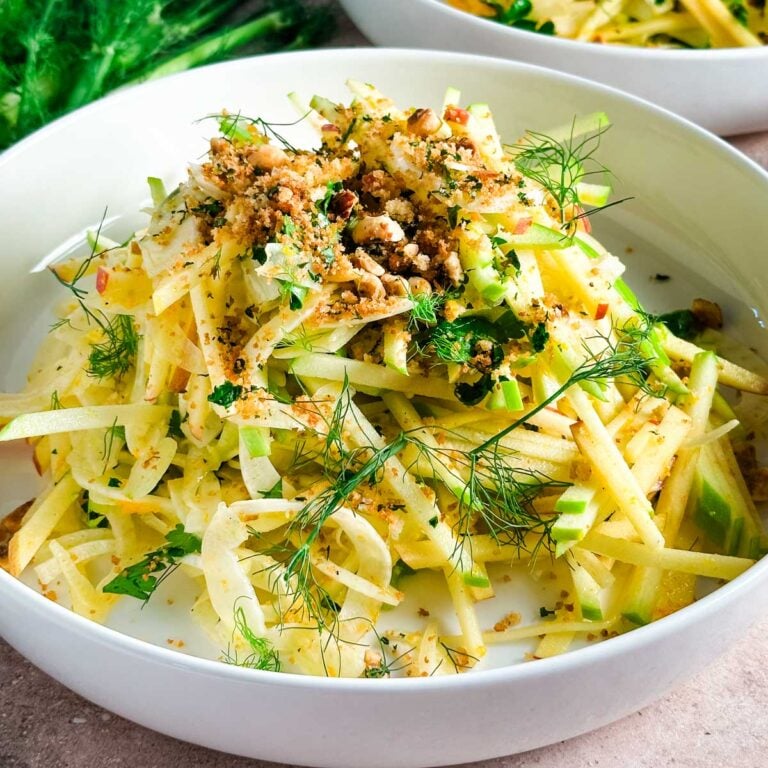 A white shallow bowl with apple fennel slaw in a high pile next to fennel fronds.