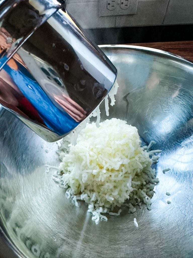Cooked and dried potatoes being passed through a ricer into a large stainless steel bowl.