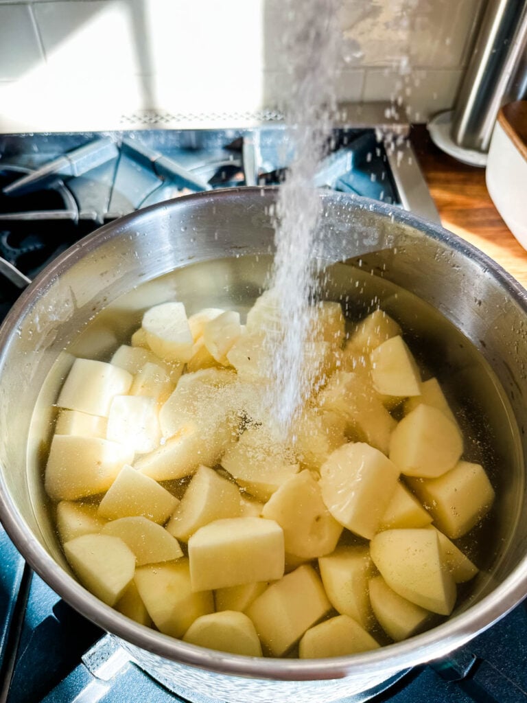 A pot of peeled and cut potatoes covered with water and a stream of kosher salt being added before cooking.
