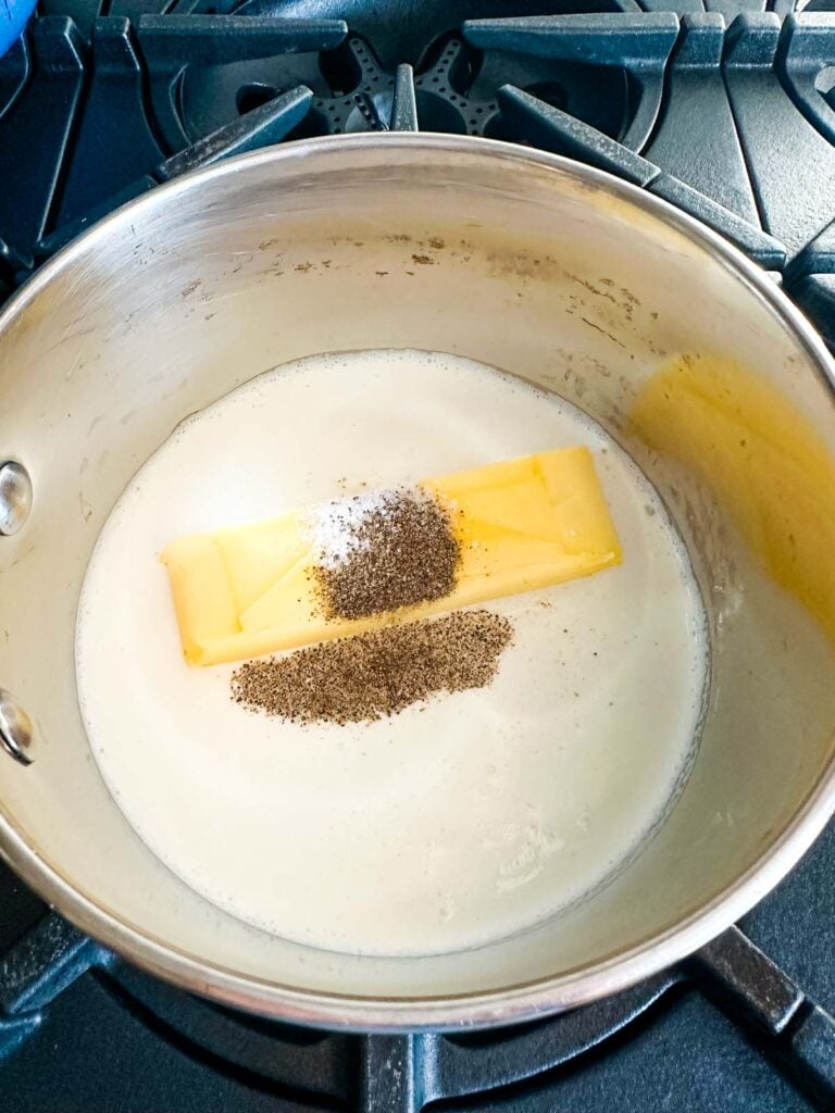 A small saucepan with heavy cream, butter, salt and pepper heating up on the stovetop.