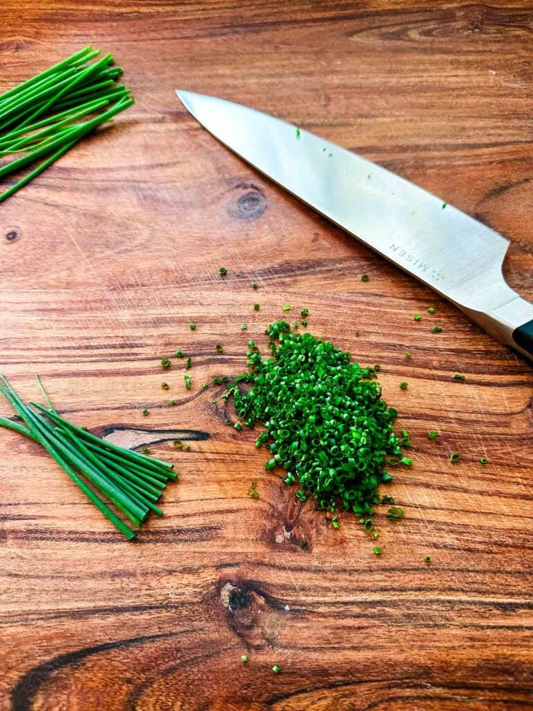 A cutting board with chopped chives next to a chef's knife.