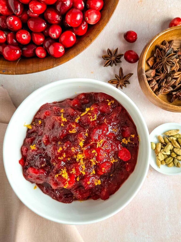 A bowl of spiced cranberry sauce next to fresh cranberries, a bowl of star anise pods and whole cardamom.