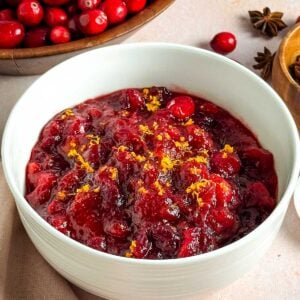 A white bowl filled with spiced cranberry sauce next to a bowl of cranberries and spices.