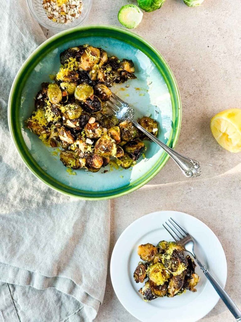 A green serving bowl of roasted Brussels sprouts next to a dish with a fork.