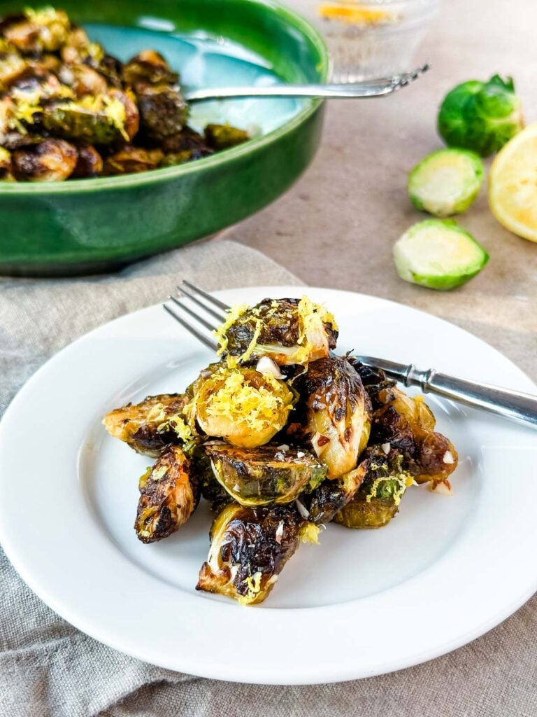 A small white plate of roasted brussels sprouts next to a green serving bowl, raw brussels sprouts, and a lemon.