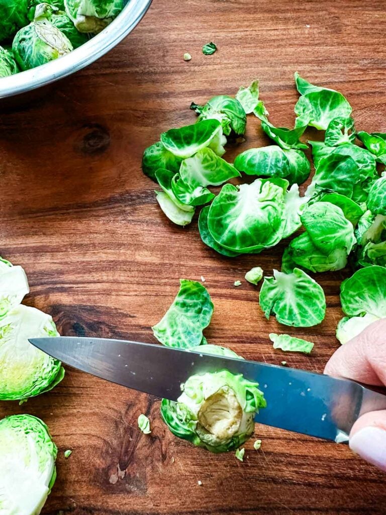 A hand with a pairing knife showing how to trim the root end of the brussels sprout and remove any damaged leaves.