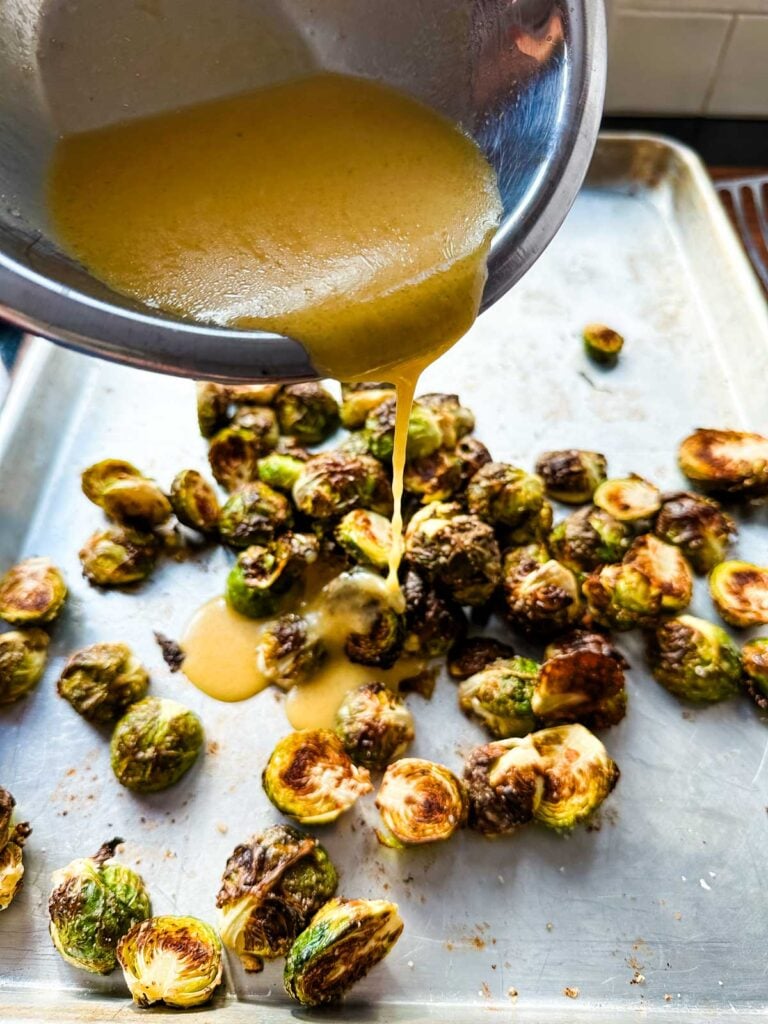 A stainless steel bowl of miso glaze being poured onto roasted brussels sprouts.
