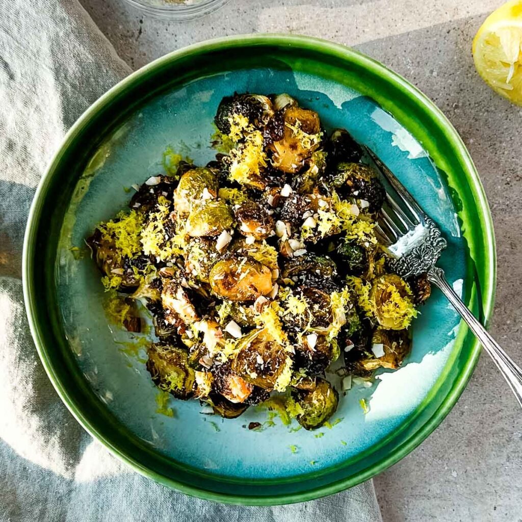 A green ceramic bowl of roasted brussels sprouts with a serving fork on a beige linen napkin.