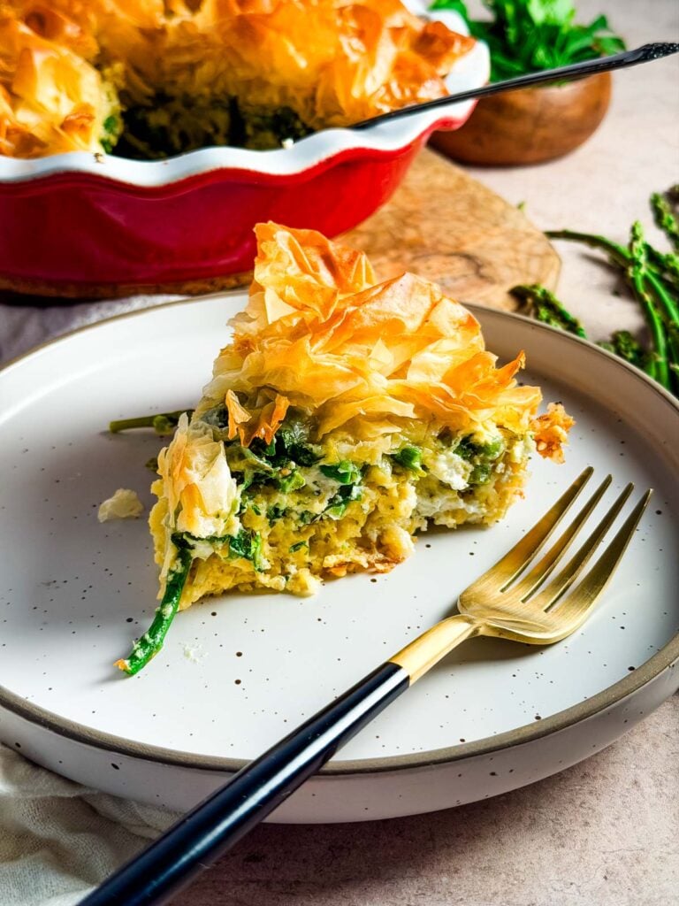 A slice of asparagus tart on a plate with a gold fork next to the red tart pan.