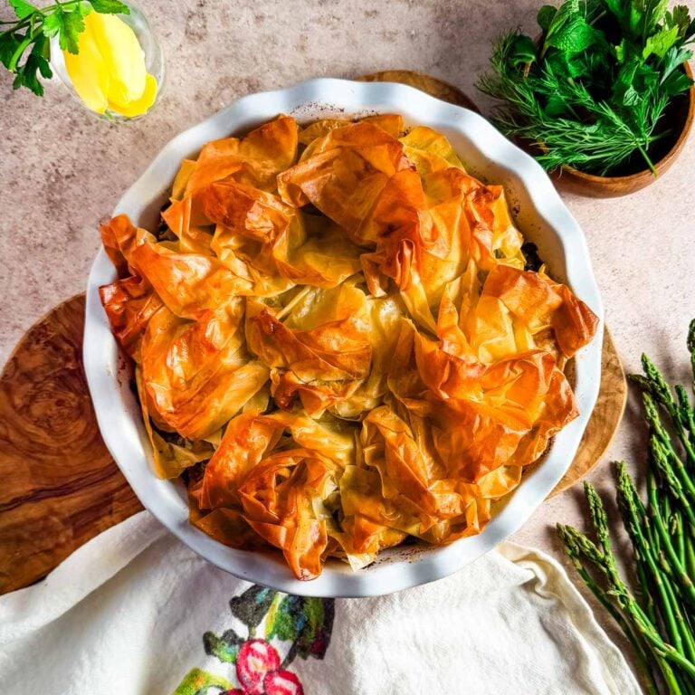 An overhead view of asparagus tart in a deep pie dish surrounded by asparagus, herbs, and a kitchen towel.