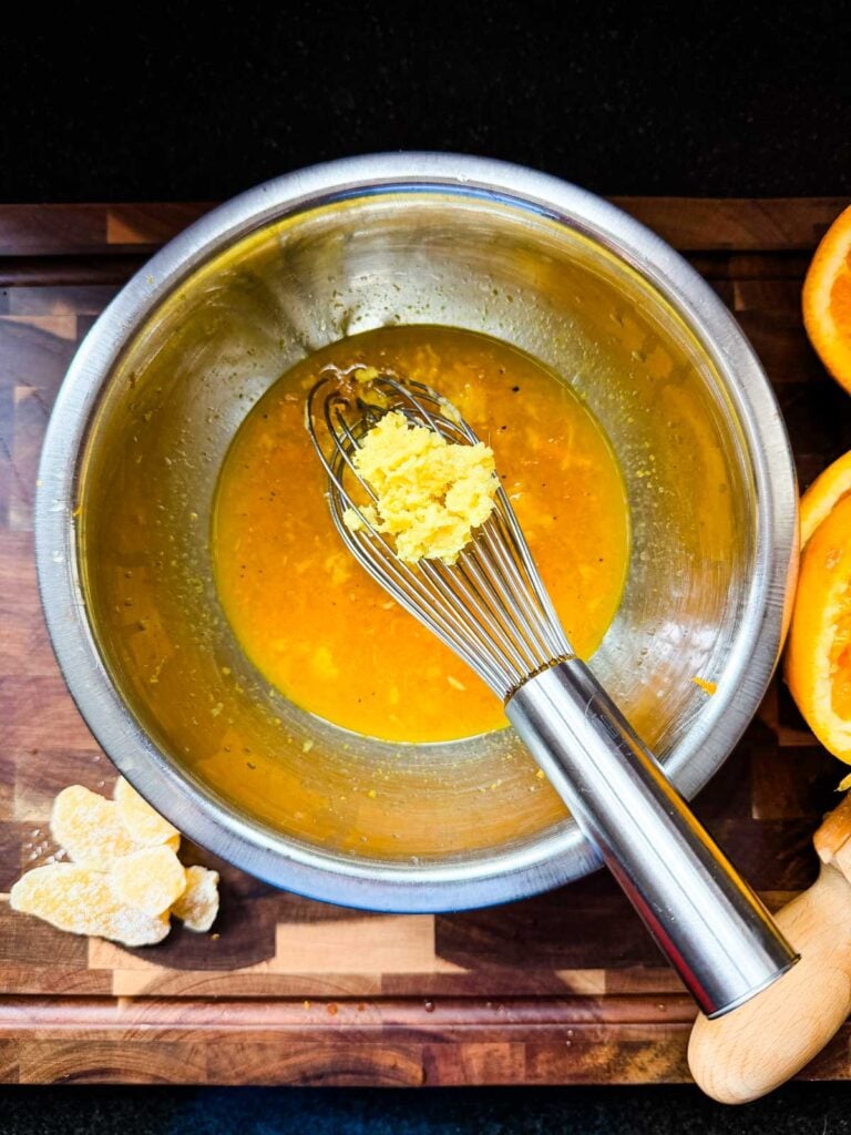 Grated ginger and garlic on a whisk in a bowl of vinaigrette on top of a cutting board.